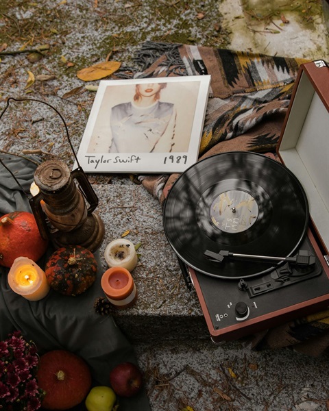 Taylor Swift's 1989 album is the at the center of this image surrounded by a fall setting with a vinyl, a pumpkin, a blanket and a lantern around it.